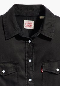 Svart denimskjorte med spiss krage, to frontlommer, knappefeste og hvite knapper. Har en rød Levi's-merkelapp inne i nakken.