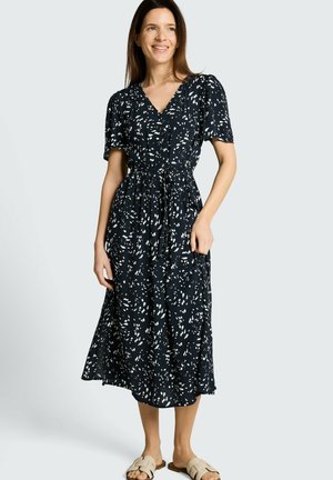 Kobieta stoi, ubrana w czarno-białą midi sukienkę w kropki z krótkimi rękawami oraz beżowe sandały wsuwane, uśmiechając się i patrząc na bok.