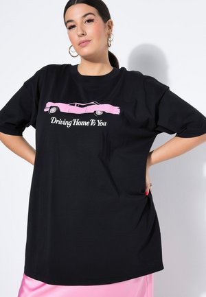 Frau mit Creolen trägt ein schwarzes oversized T-Shirt mit pinkem Auto-Grafik und dem Schriftzug „Driving Home To You“, kombiniert mit einem pinken Rock.