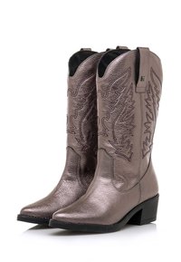 Botas vaqueras de bronce metálico con puntera puntiaguda, cuero texturizado y costuras decorativas, que presentan detalles recortados en la parte superior.