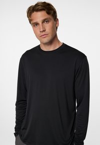 Mustapaita pitkillä hihoilla, valmistettu pehmeästä kankaasta, jossa on pyöreä pääntie ja huomaamaton logo-details rinnassa. Sileä materiaali, jossa istuvat hihat.