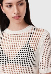 Hvid strikket sweater med åben mesh-design, korte ærmer og rund halsudskæring, lagd ovenpå en sort bh. Har en tekstureret overflade.