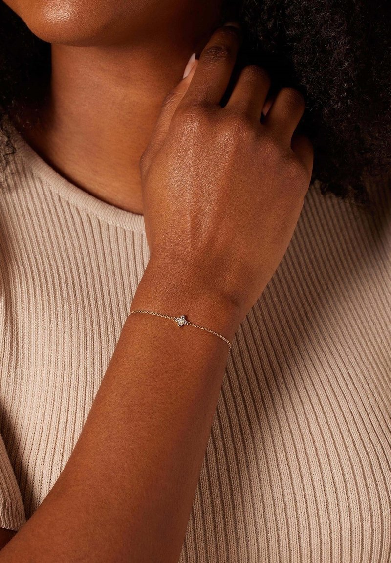 Zilveren schakelarmband met een centraal decoratief element. Gemaakt voor een hand met een gladde, donkere huidtone. De achtergrond toont een ribbelstof.
