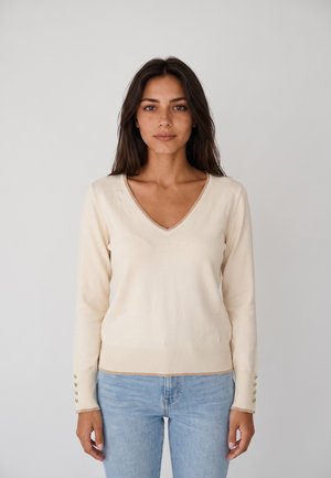 Donna con capelli lunghi e scuri che indossa un maglione crema con scollo a V e bottoni sulle maniche e jeans azzurri, in piedi davanti a uno sfondo semplice.