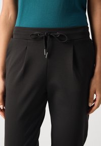 Persona che indossa pantaloni neri con il cordino e una maglietta teal, inquadratura ravvicinata dalla vita a metà coscia, mani rilassate ai fianchi.