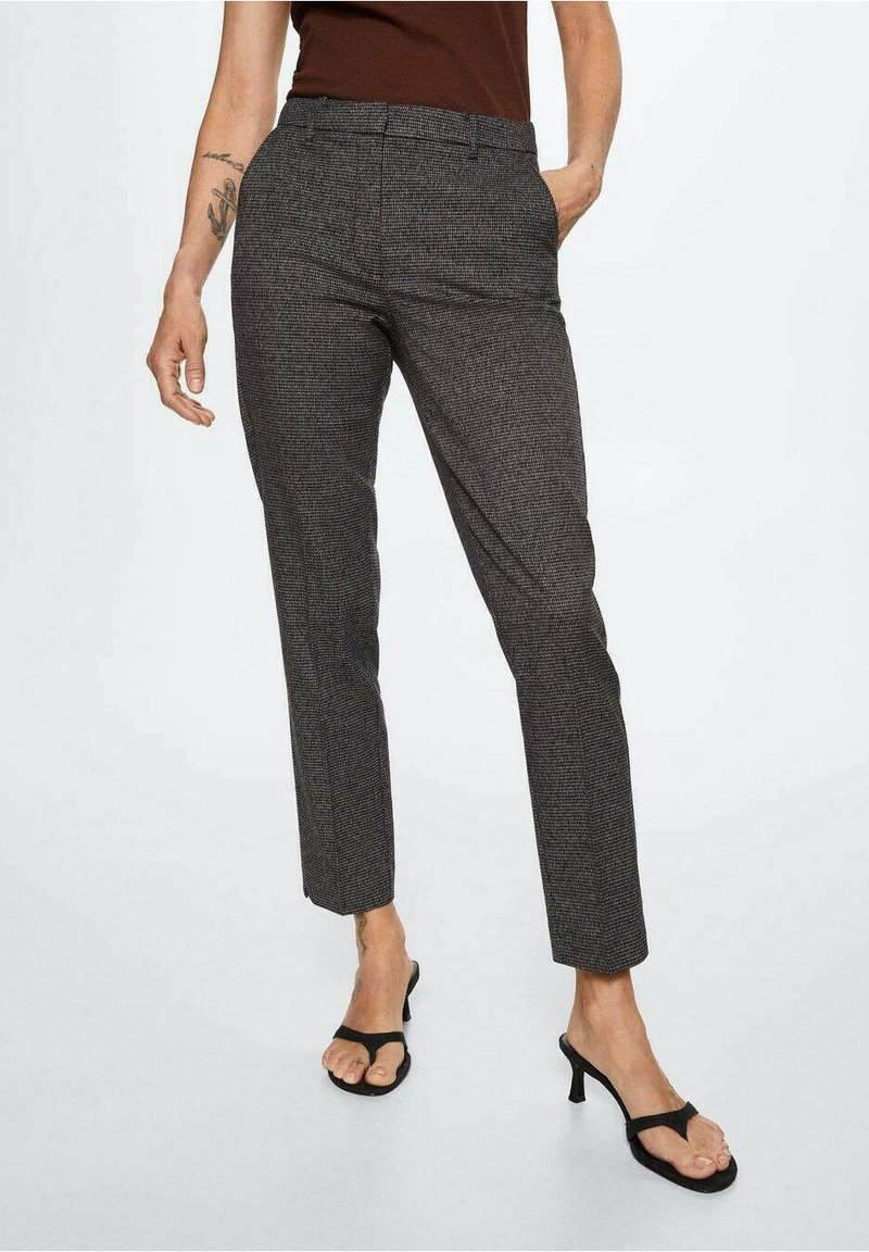 Femme portant un pantalon gris texturé ajusté avec poches et des sandales à talons noires, debout devant un fond uni.