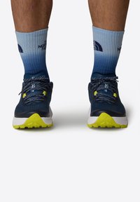 Modré sportovní boty s reflexními tkaničkami, žlutými akcenty a texturovanou podrážkou, spárované s gradientovými modrými ponožkami se logem.