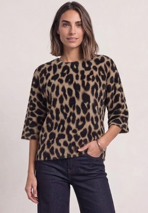 Femme aux cheveux mi-longs porte un pull à imprimé léopard et un jean foncé, avec une main dans la poche et un bracelet argenté au poignet.