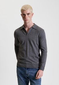 Graues langärmeliges Polohemd mit Kragen und einer Dreiknopf-Leiste. Weicher Stoff mit gerippten Bündchen und Saum, kombiniert mit blauen Jeans.