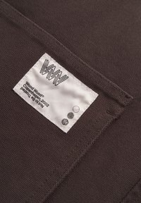 Rjava tekstilna površina v close-upu, ki prikazuje belo oznako z črnim logotipom in ikonami, prišito ob robu. Tkanina ima teksturirano površino.