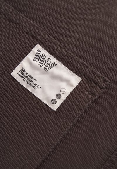 Prim-plan cu un material textil maroniu ce prezintă o etichetă albă cu un logo și icoane negre, coasă la margine. Țesătura are o suprafață texturată.