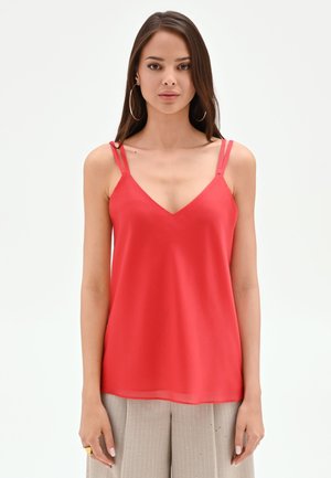 Femme aux cheveux bruns raides portant de grandes boucles d'oreilles créoles, un haut rouge sans manches à col en V avec des bretelles doubles et un pantalon beige à fines rayures.