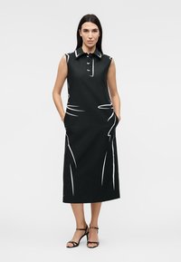 Femme portant une robe midi noire sans manches avec des motifs abstraits blancs en lignes, col boutonné, poches et talons noirs à brides, debout avec assurance.