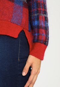 Nahaufnahme einer Hand einer Person, die an ihrer Seite ruht, während sie einen rot-blauen karierten Strickpullover und dunkelblaue Jeans trägt.