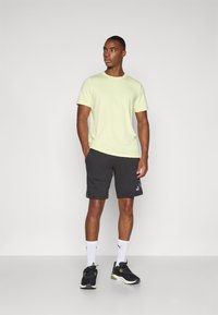 Lys gul atletisk skjorte, mørke shorts med sidelommer, sorte sko, hvide sokker. Enkel design, glat struktur og logo detaljer synlige.