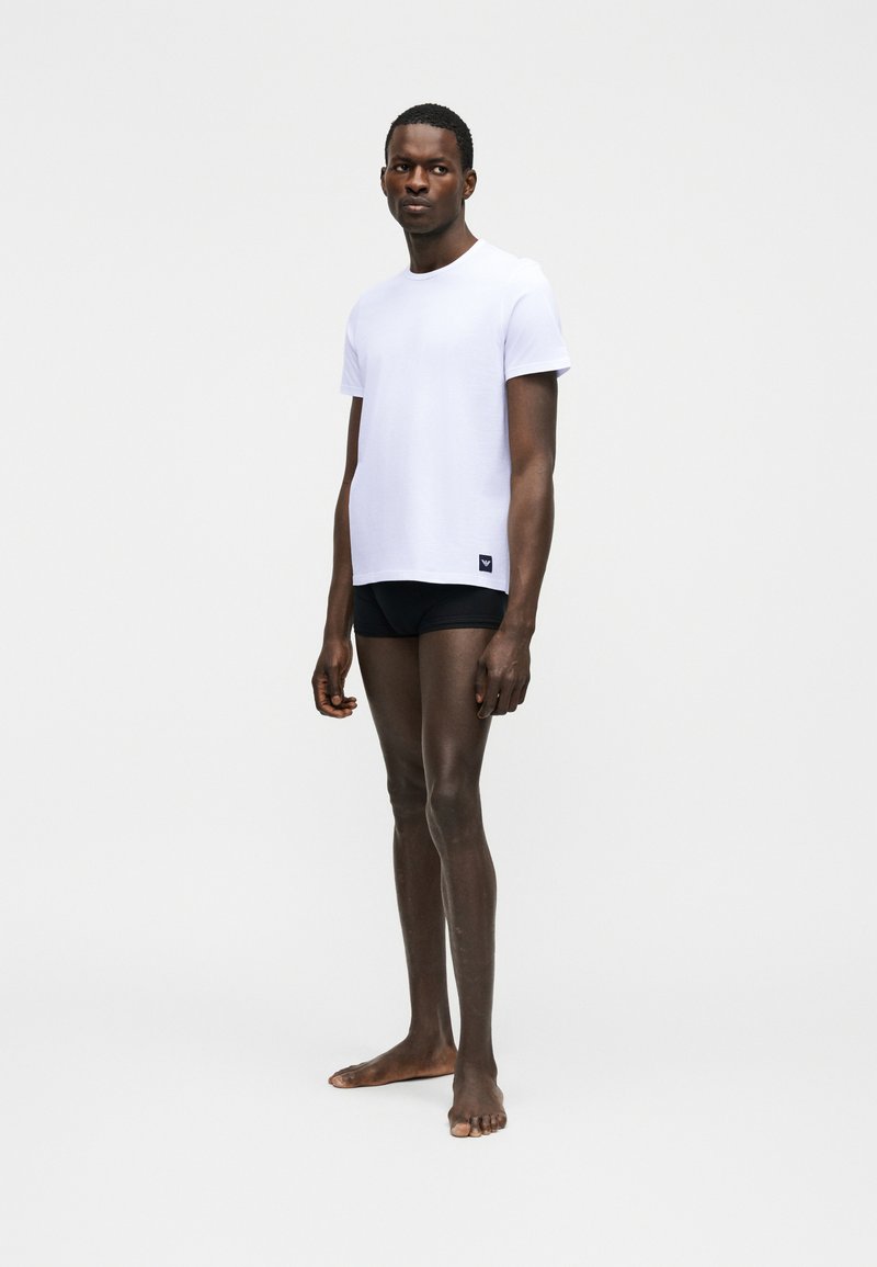 Άνδρας με σκούρο δέρμα, όρθιος και ξυπόλητος, φοράει απλό λευκό κοντομάνικο μπλουζάκι και μαύρα εσώρουχα τύπου μπόξερ, μπροστά σε απλό φωτεινό φόντο.