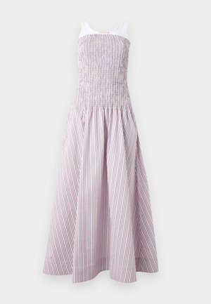 Pitkähiuksinen tummahiuksinen nainen, joka pukeutuu hihattomaan vaaleanpunaiseen ja valkoiseen raidalliseen midimekkoon ja beigen värisiin terävälokkeisiin tasapohjakenkiin, seisoo käsi lantiolla.