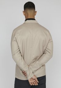 B beige spalvos švarkas su struktūrizuotu dizainu, ilgomis rankovėmis ir apykakle. Audinys atrodo lygus ir turi subtilų blizgesį.