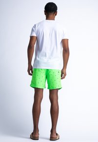 Męska biała koszulka, widok z tyłu, w połączeniu z neonowozielonymi szortami kąpielowymi z czarnym wzorem w trójkąty. Gładki materiał i swobodny styl.