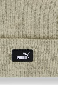 Beanie lavorato a maglia beige con risvolto e una toppa rettangolare nera con il logo bianco "PUMA" e una silhouette di un gatto che salta.