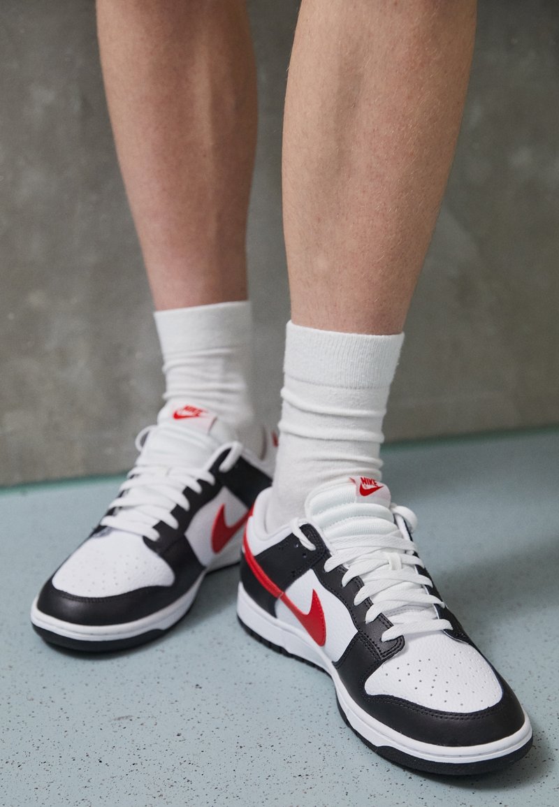 Jambes portant des chaussettes blanches montantes et des baskets Nike noires, blanches et rouges sur un sol bleu clair tacheté.