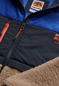 Gevoerde jas van blauw en zwart nylon, met oranje ritsen en een tan gekruiste fleece sectie aan de onderkant. Merklogo zichtbaar.