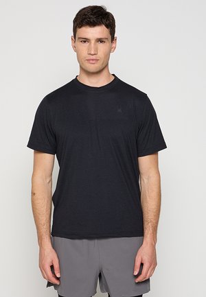 Młody mężczyzna z krótkimi ciemnymi włosami, ubrany w gładką czarną koszulkę i szare krótkie spodenki, stoi zwrócony przodem na jasnym tle.