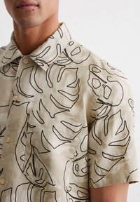 Beige Leinenhemd mit kurzen Ärmeln, das schwarze botanische Linienmuster, einen Kragen und eine Brusttasche aufweist. Die Knöpfe sind hell gefärbt.