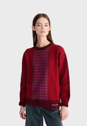 Jeune femme portant un pull rouge et violet à motifs et un pantalon bleu foncé, debout devant un fond gris clair uni.