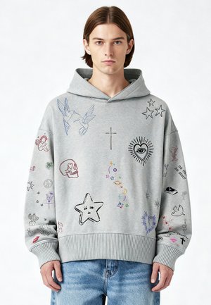 Jeune homme portant un sweat à capuche gris orné de dessins colorés, comprenant des oiseaux, des étoiles, un crâne, un cœur avec un œil, et divers petits symboles, assorti à un jean bleu.
