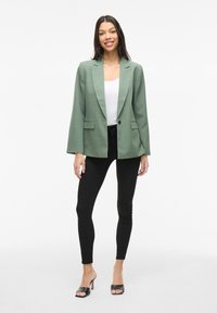 Blazer verde con finitura satinata, dotato di rever a notched, due tasche frontali e chiusura con un solo bottone, abbinato a leggings neri.