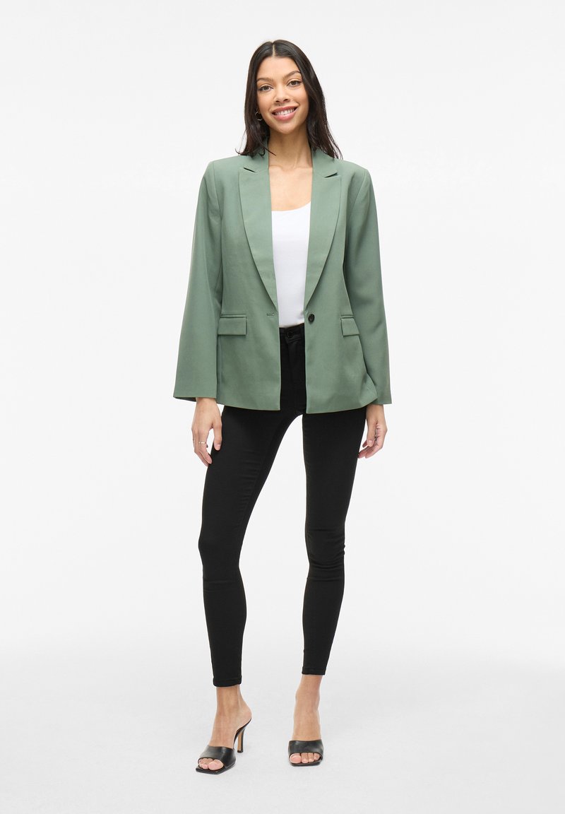 Blazer vert avec une finition satinée, doté d'un col cranté, de deux poches avant et d'une fermeture à un bouton, associé à des leggings noirs.