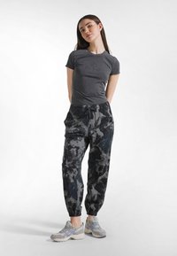 Harmaa istuva t-paita, jossa on hillitty logo, yhdistetty mustavalkoisiin kukkakuviollisiin collegehousuihin ja vaaleisiin lenkkareihin, joissa on violetteja yksityiskohtia.