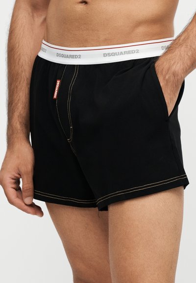 Ανδρικός κορμός που φοράει μαύρα boxer Dsquared2 με λευκή ελαστική μέση και εμφανή λογότυπα της μάρκας, με το ένα χέρι στην τσέπη.