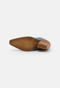 Zapato de tela azul con un tacón de madera medio y una suela marrón texturizada. Presenta un puntera puntiaguda y una marca grabada en el lateral.