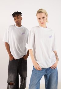 Λευκά oversized t-shirts με πορφυρό κεντητό σχέδιο στο αριστερό στήθος, φορεμένα με σκούρα σκισμένα τζιν και ανοιχτόχρωμα φαρδιά τζιν.