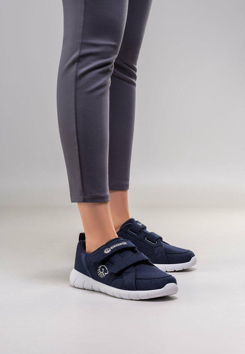 Ben in donkergrijze leggings met marineblauwe Giesswein sneakers met witte zolen en klittenbandsluitingen, staand op een lichtgrijze vloer.