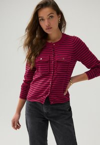 Cardigan fuchsia avec des rayures horizontales noires, présentant deux poches avant et un col rond. Associé à un jean gris foncé.