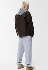 Giacca marrone oversize con cappuccio a quadri grigio, abbinata a pantaloni della tuta grigi larghi. Dotata di tasche laterali e vestibilità rilassata.