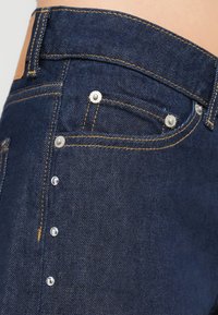 Σκούρες μπλε τζιν παντελόνες διαθέτουν ασημί αξεσουάρ, αντίθετη πορτοκαλί ραφή και δύο μπροστινές τσέπες με διακοσμητικά καρφιά.