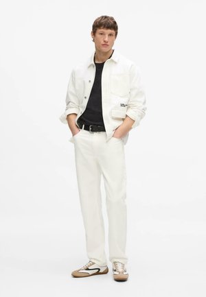 Młody mężczyzna ubrany w białą kurtkę i spodnie na czarnej koszuli, stoi z rękami w kieszeniach, beżowe i białe sneakersy na jednolitym białym tle.