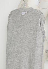 Grå ärmlös tröja i stickad tyg, med rund halsringning och subtil textur, med en etikett synlig på baksidan.