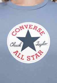 Ανοιχτό μπλε μπλουζάκι με στρογγυλό λευκό λογότυπο που περιλαμβάνει ένα σκούρο μπλε αστέρι και κόκκινο κείμενο που γράφει "Converse Chuck Taylor All Star".