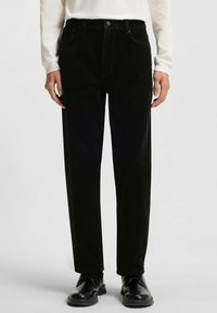 Pantaloni di velluto a coste neri con design a gamba dritta, dotati di tasche frontali e chiusura con bottone, abbinati a scarpe nere.
