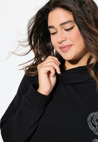 Felpa nera con collo a cowl e maniche lunghe, caratterizzata da un motivo di accento argentato. Tessuto morbido e vestibilità comoda.