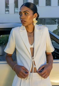 Blazer blanc à manches courtes avec un fermoir à cravate, superposé à un bralette en dentelle, associé à une jupe de couleur claire assortie. Boucles d'oreilles en fleurs embellies.