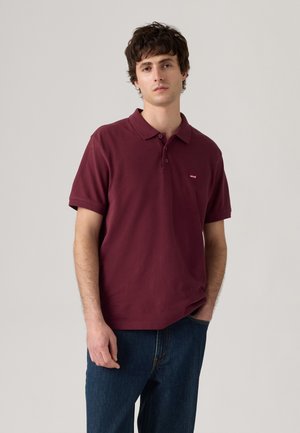 Tricou polo burgundy, cu mânecă scurtă, guler cu trei butoane, detaliu mic cu logo pe pieptul stâng, combinat cu blugi albaștri.