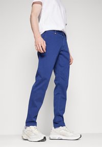 Blå bomullshangarer med rak passform och sidofickor, i kombination med vita sneakers. Har gylf med dragkedja och orange accent på midjebandet.