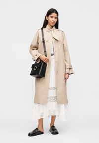 Žena v béžovom trenčkote cez biele čipkované šaty, nesie čiernu kabelku, stojí pred jednoduchým svetlým pozadím.