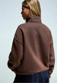 Bruine fleece sweatshirt met hoge kraag, losse pasvorm, verlaagde schouders en zachte textuur, gecombineerd met donkerdenim jeans.